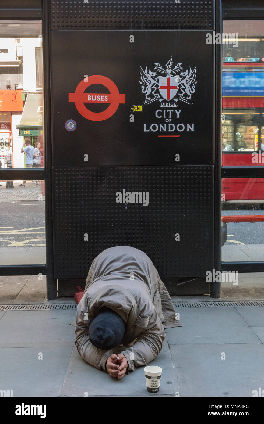 A homeless person begging next to a bus shelter in the city of london financial district. Chalk and cheese and contrasts. Lucky and unlucky situations Stock Photo