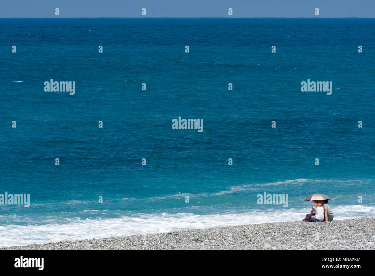 Back view of lovely couple with parasol sitting on beautiful pebble beach and enjoying sunny day, overlooking the sea, Chisingtan, Hualien, Taiwan Stock Photo
