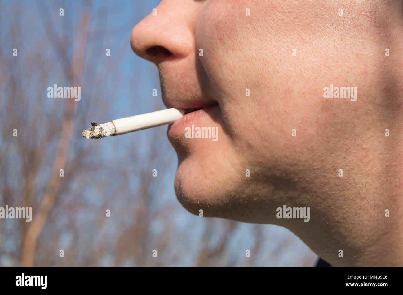 how to hold a cigarette in mouth Young handsome man holding a cigarette in his mouth. Stands against the  blue sky Stock Photo