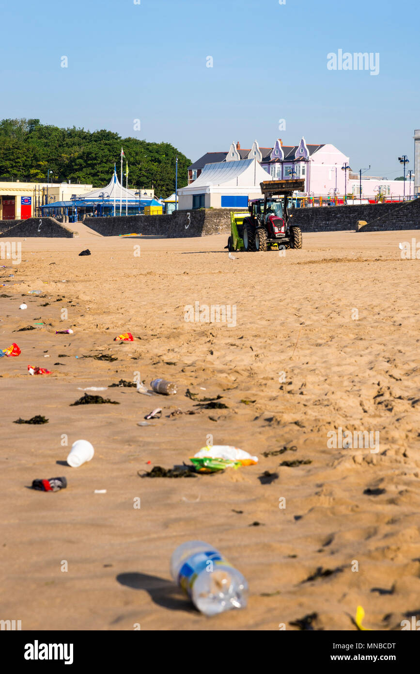 Foreground litter on quiet sandy beach at tourist resort early on a bright sunny summer morning being cleaned by background tractor with surf rake. Stock Photo
