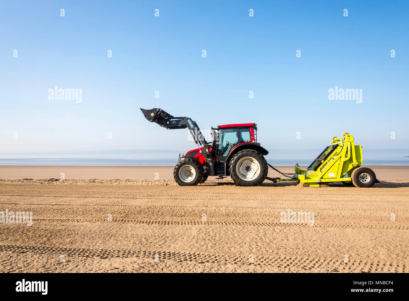 Whitmore Bay beach at Barry Island, Wales,early on a bright sunny summer morning being cleaned of litter by a red Tractor pulling a Barber Surf Rake. Stock Photo