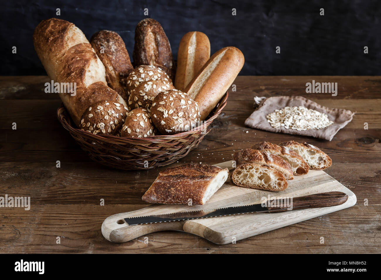 An assortment of bread in a basket and a baguette on the cutting board ...