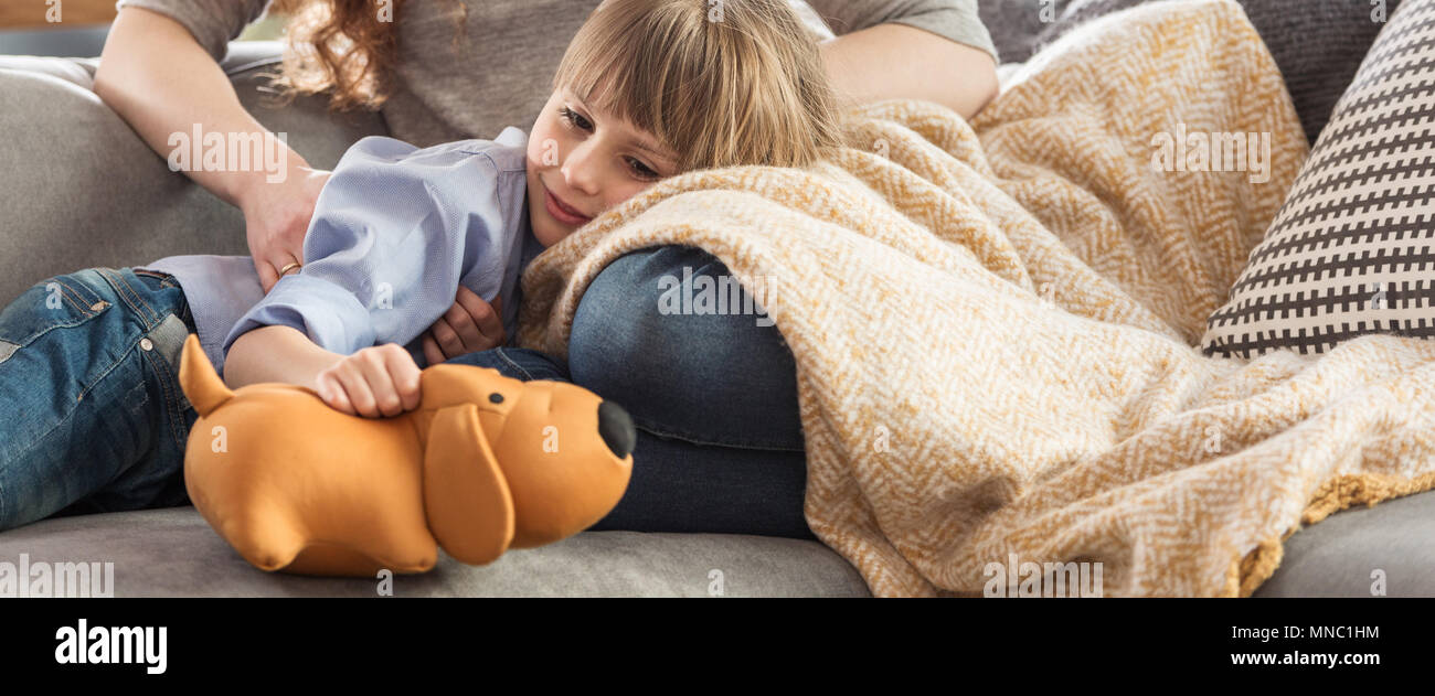 Panorama of smiling boy playing with yellow plush dog while lying with his mother on a couch Stock Photo