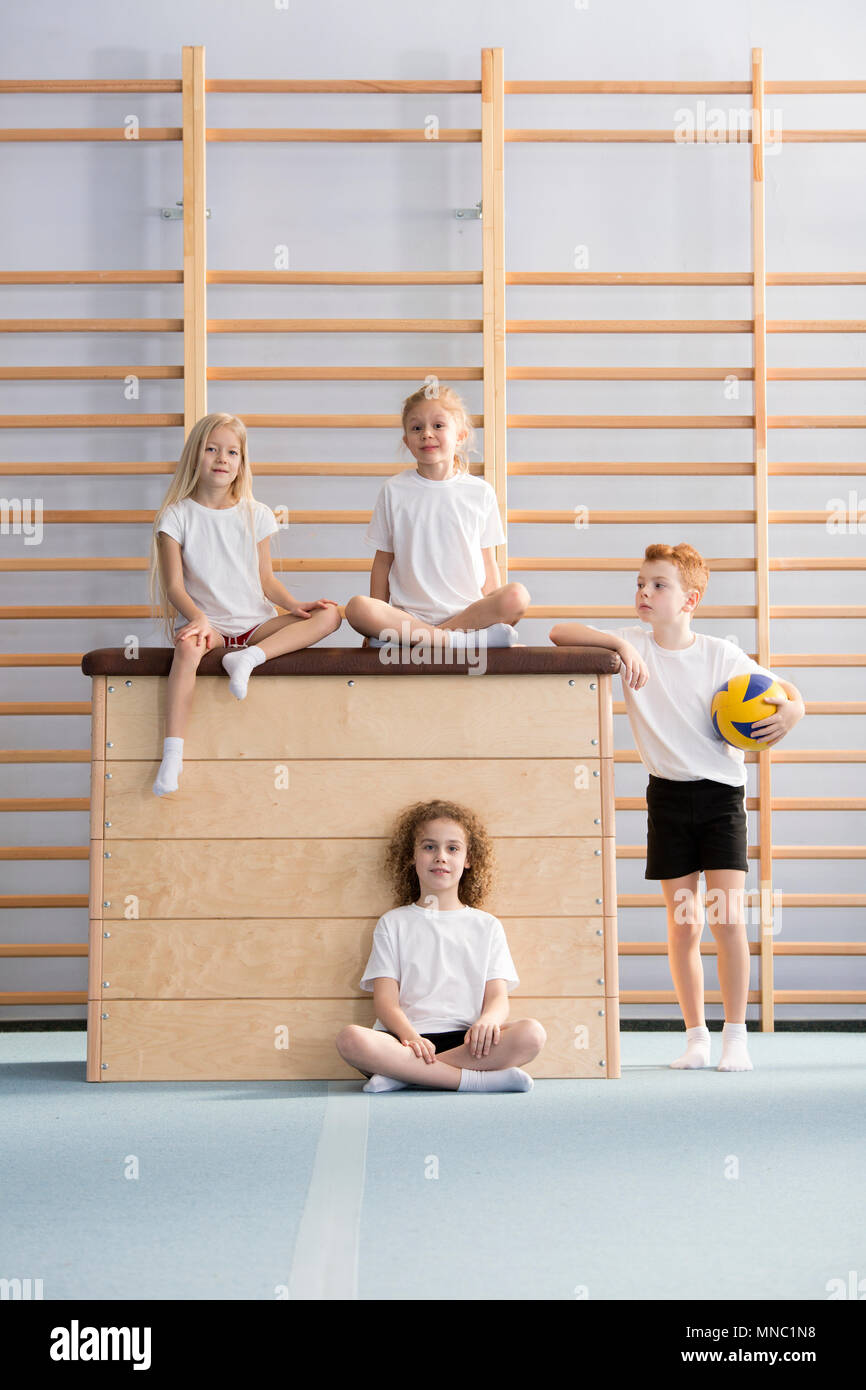Primary school girls sitting on top of the vaulting box and a boy holding a volleyball during a game break at gym class Stock Photo