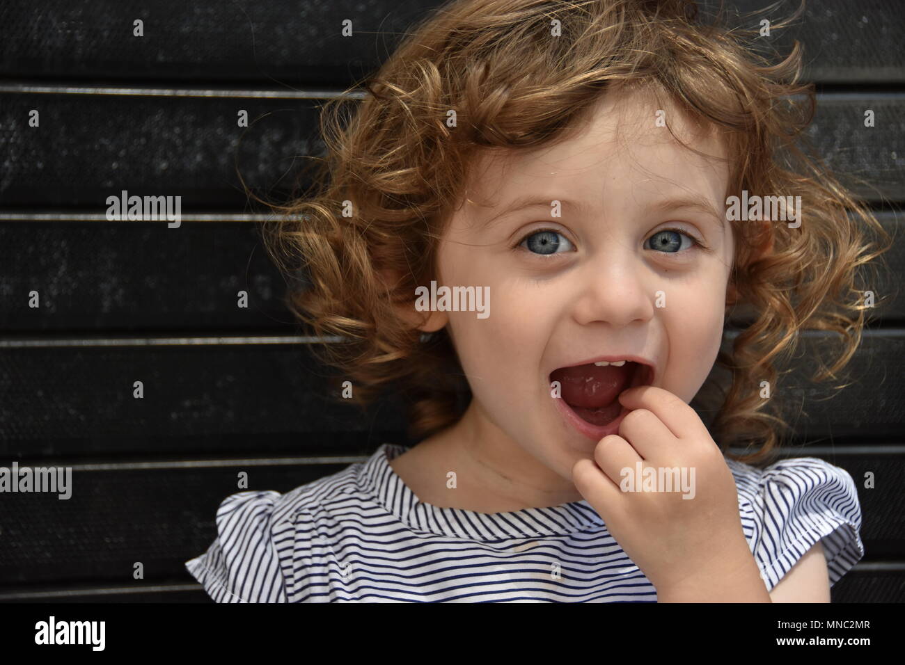 Angelic young curly haired girl points inside her open mouth with her finger sitting against a black brick wall in new york city Stock Photo