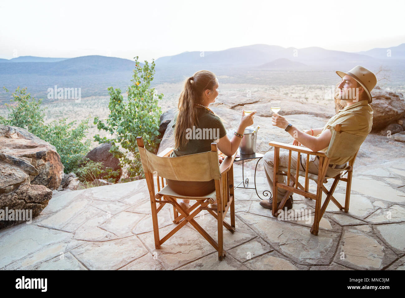 Romantic couple on safari vacation sitting outdoor at lodge enjoying stunning views over national park with glass of sundowner wine Stock Photo