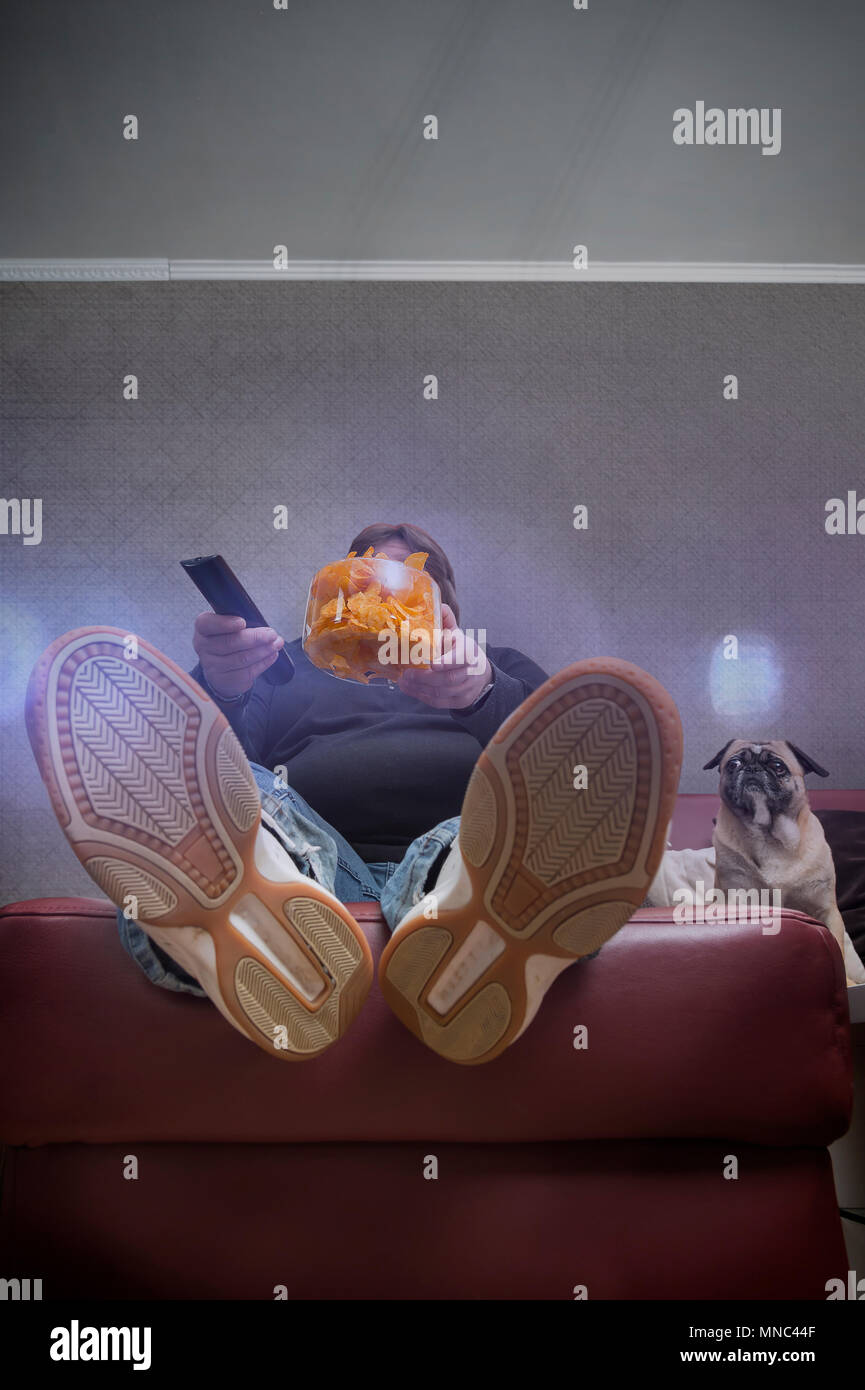 A man eats chips in front of the TV. His dog is also watching the TV. The man has a big belly. Plenty of copy space above. Worms eye view from a livin Stock Photo