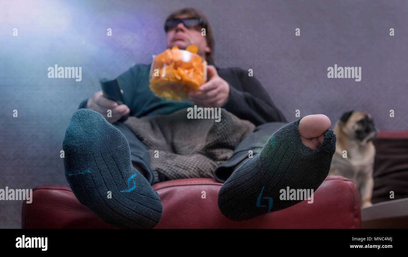 A man eats chips in front of the TV. His dog is sitting next to him on the couch. The man has a hole in the stockings and a 3 D glasses put on. Frog p Stock Photo