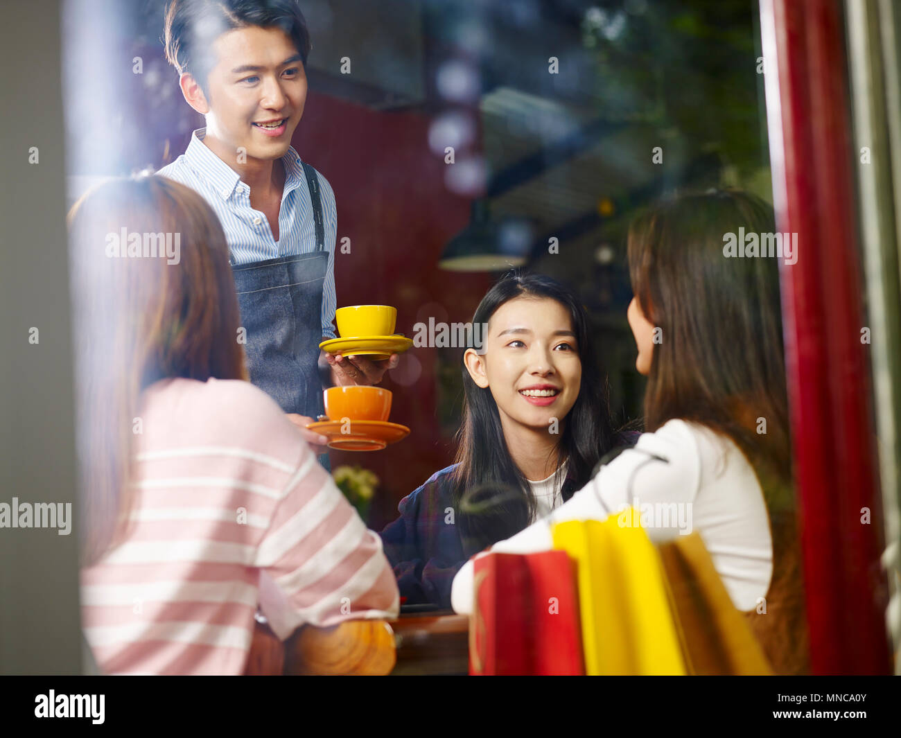 young asian waiter serving female customers in coffee shop, shot through window glass. Stock Photo