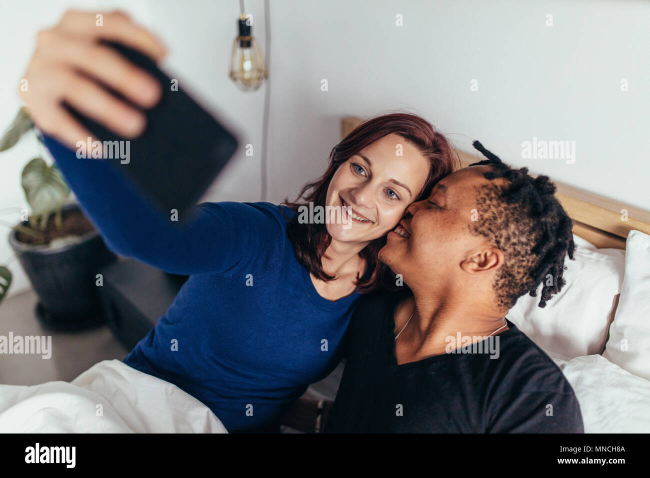 Romantic multiracial couple lying together on bed and taking selfie. Smiling man kissing on the cheek of woman taking selfie with mobile phone. Stock Photo