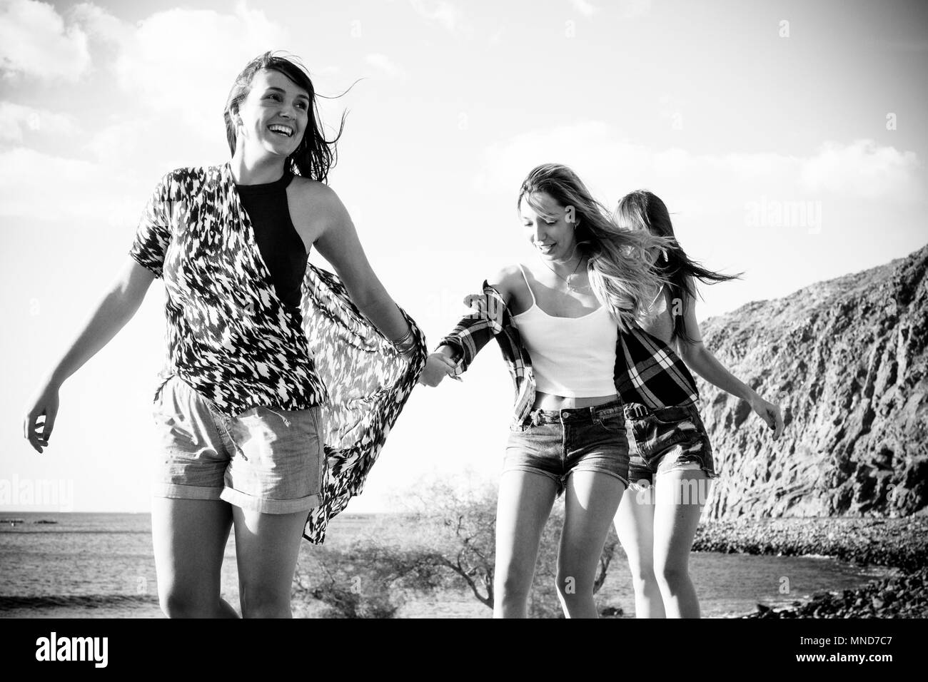Three young women friends walk together on a wall near the beach in Tenerife. Enjoy the vacation under a blue sky with yellow sun. Hands by hands. Stock Photo