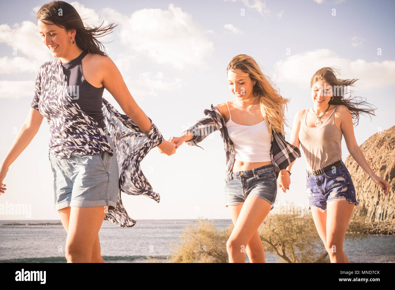 Three young women friends walk together on a wall near the beach in Tenerife. Enjoy the vacation under a blue sky with yellow sun. Hands by hands. Stock Photo