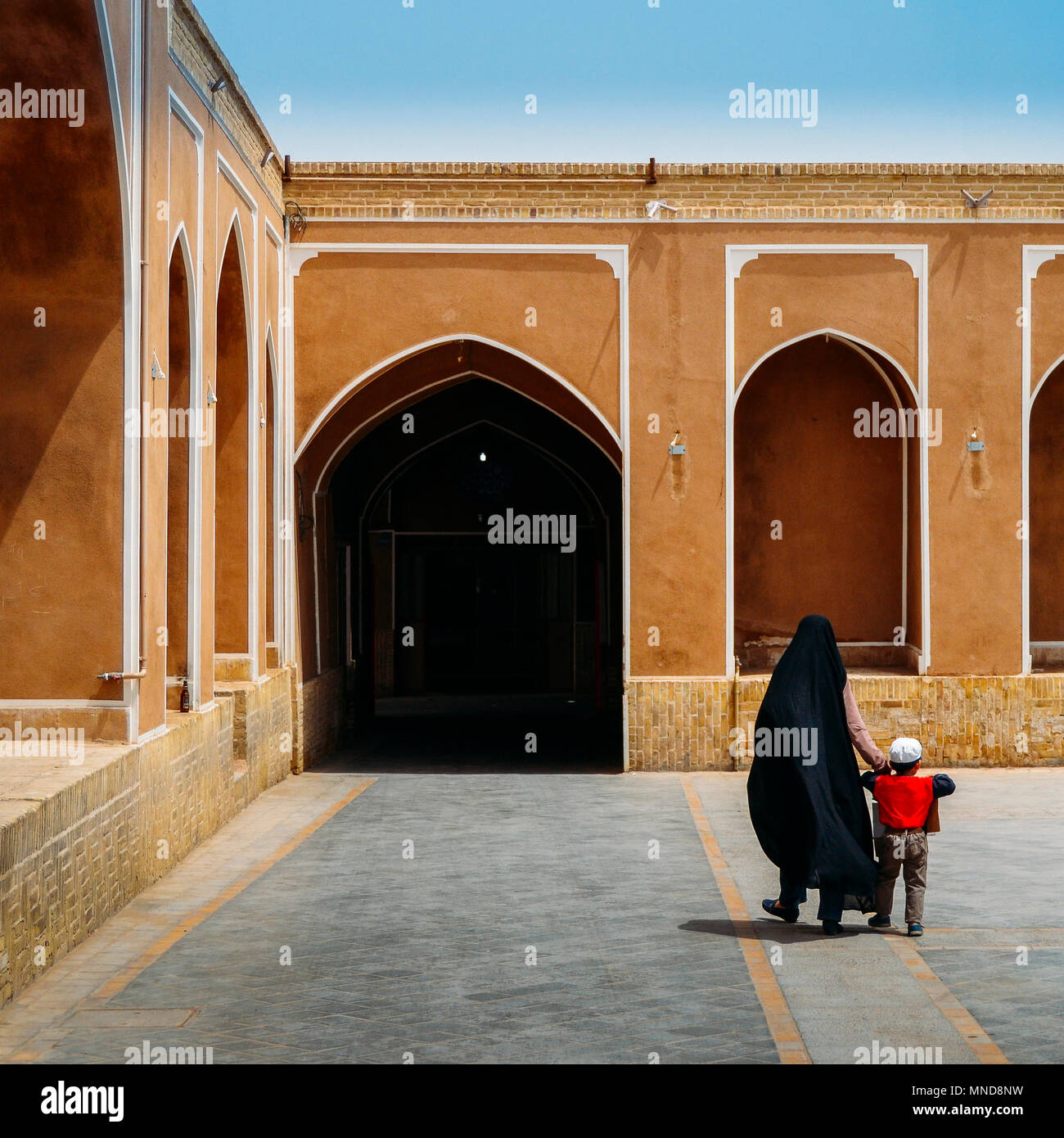 Unidentifiable woman wearing a traditional black dress in Iran, known as a chador, holds the hand of a young boy. Islamic architecture in background with ancient arches Stock Photo
