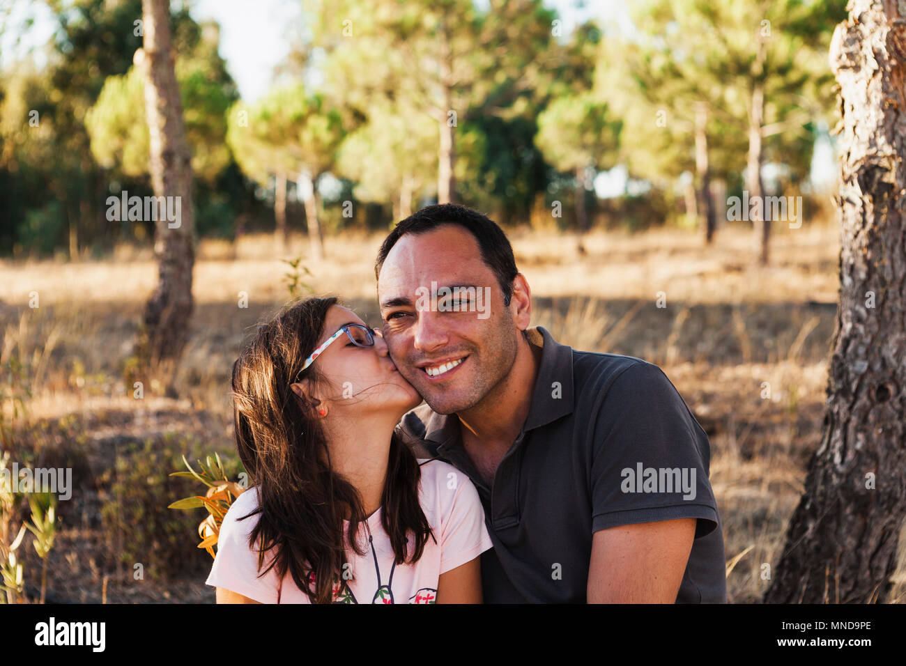 Girl kissing happy father sitting by tree trunk in forest Stock Photo
