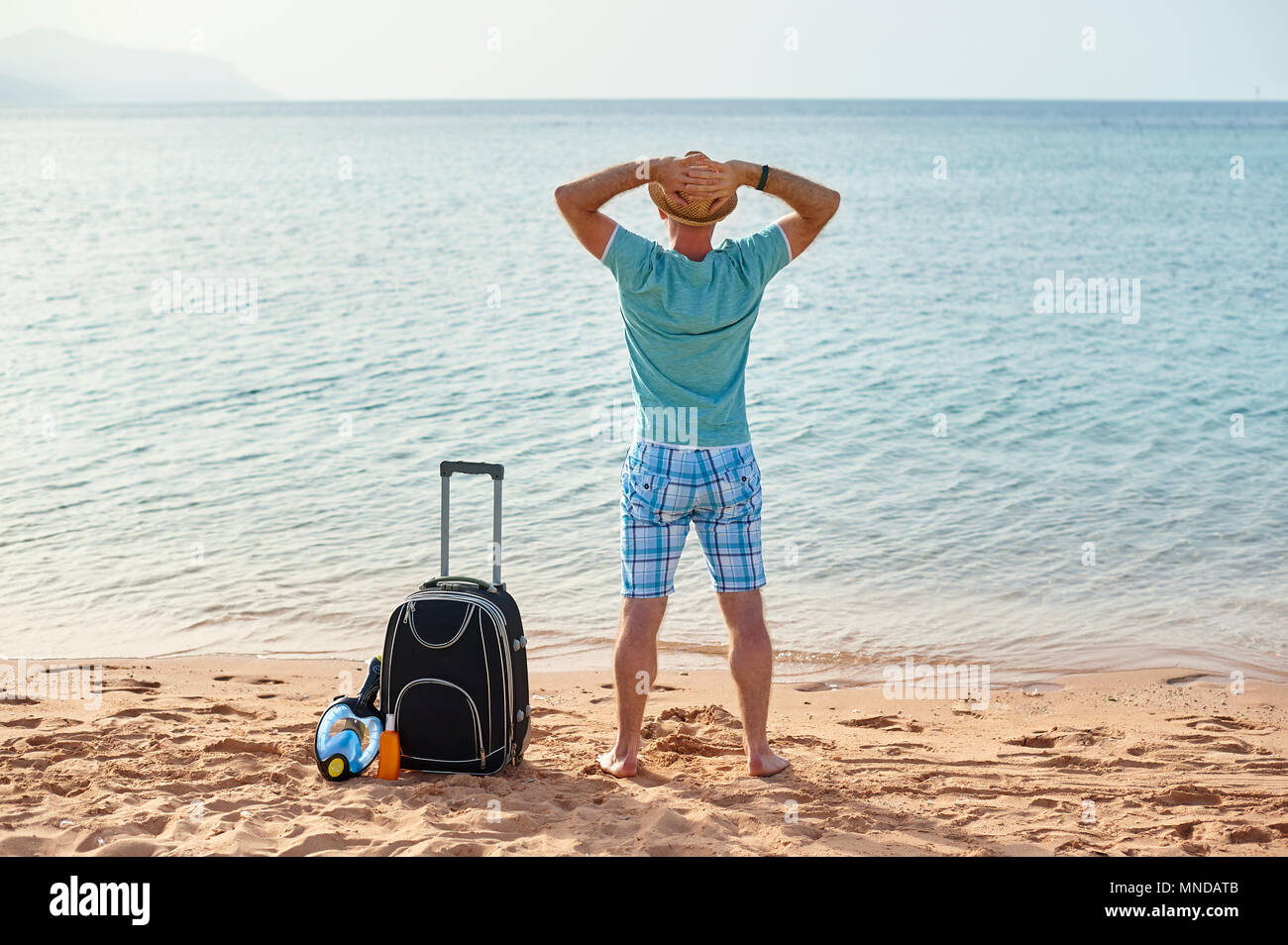 Man tourist in summer clothes with a suitcase in his hand, looking at the sea on the beach, concept of time to travel Stock Photo
