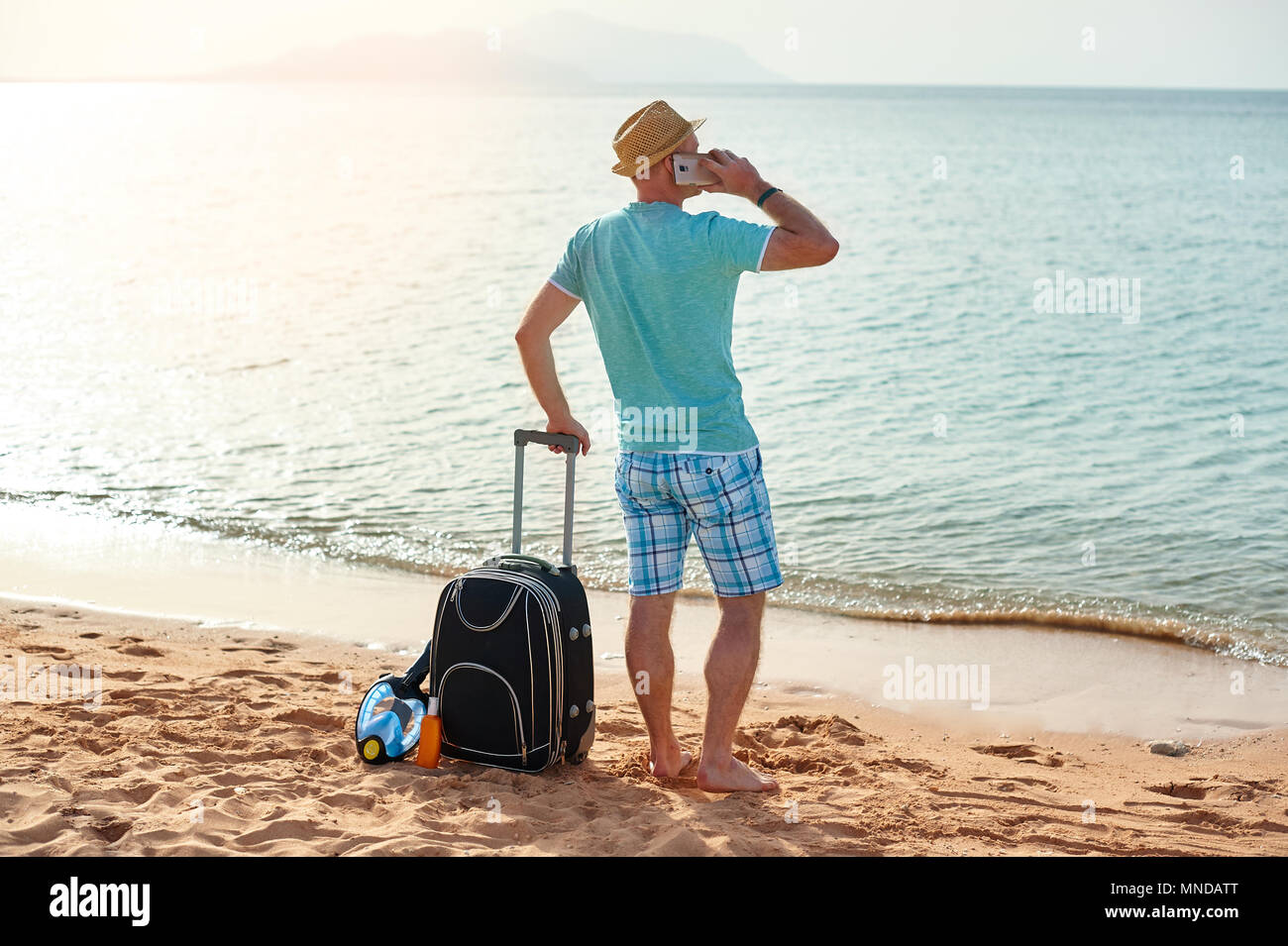 Full length portrait of happy man with suitcase and mobile phone standing on beach freelancer on a sunny day on vacation Stock Photo