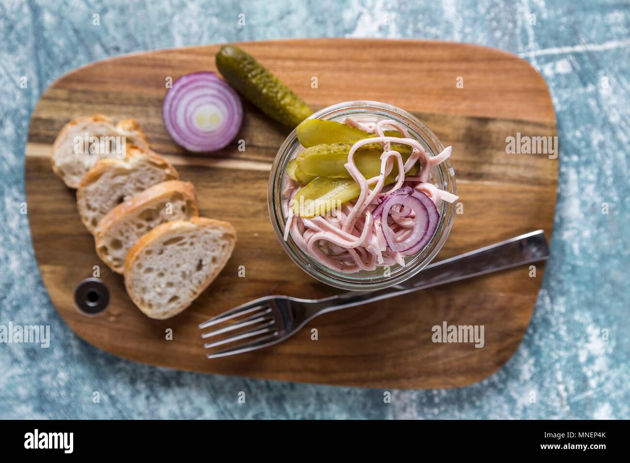 A sausage, red onion and gherkin salad in a glass on a wooden board, with baguette slices Stock Photo