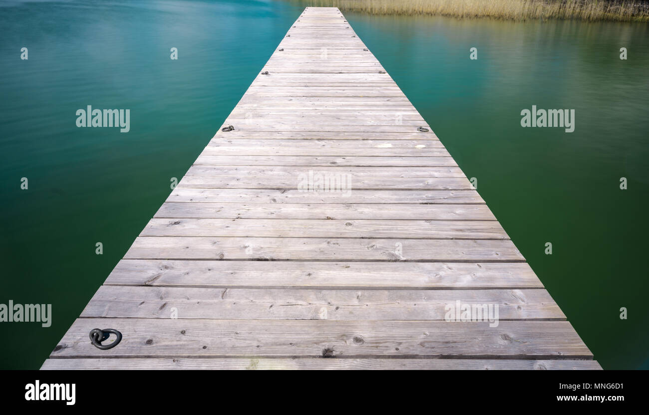 wooden pier jutting out into a turquoise and blue lake with golden reeds in the background Stock Photo