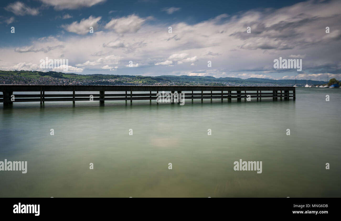 wooden pier on lake Zurich with rolling hills mountain landscape and sailboats in the background Stock Photo