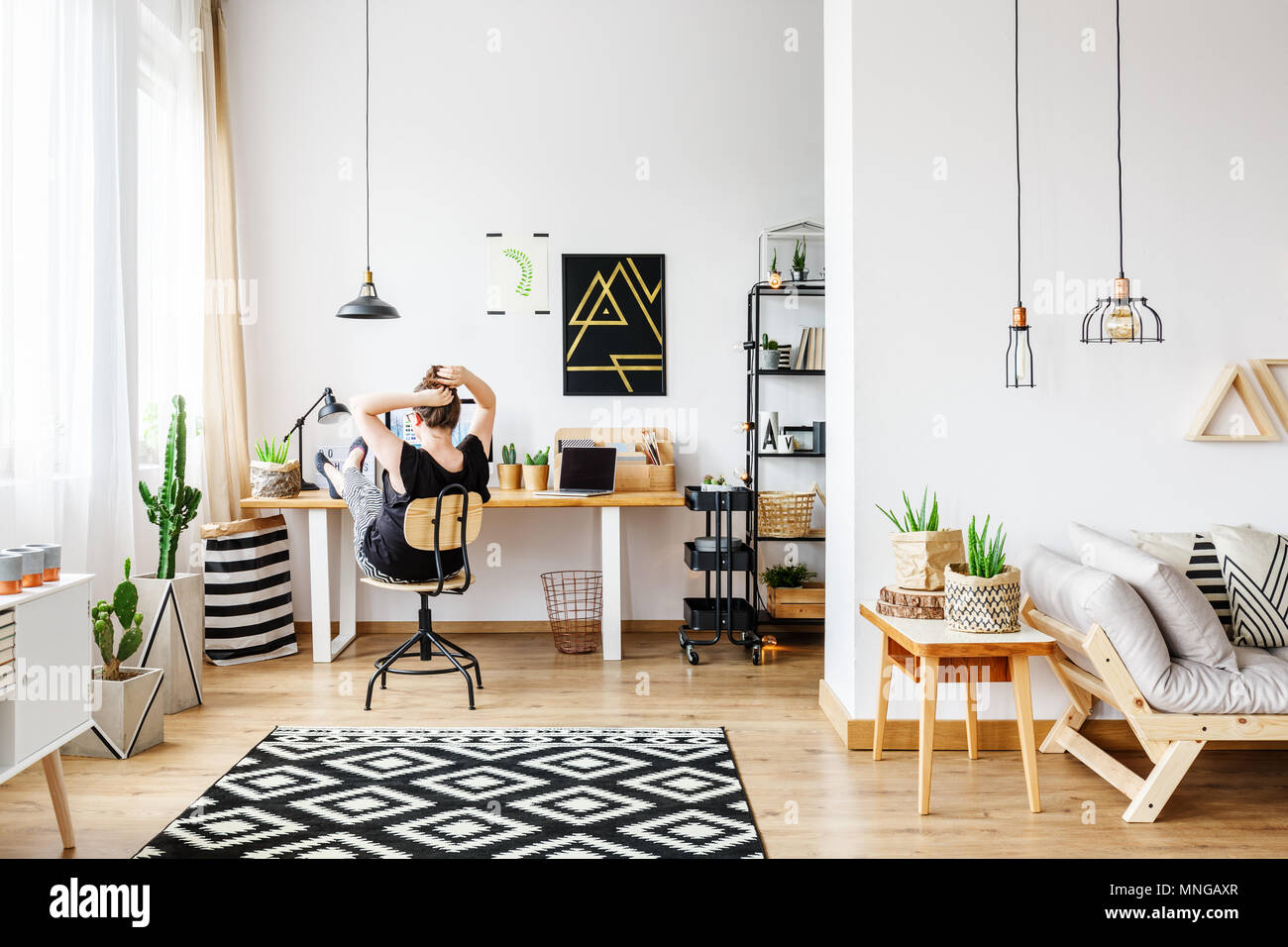 Young freelance woman working remotely at designer home in cozy white office room with pendant lamps, white wall, big window, comfy couch and aloe pla Stock Photo