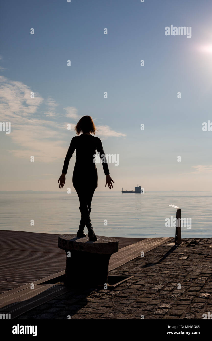 Back view of dancing young beautiful fit woman with short hair on docking pole in Thessaloniki, Greece. Rear view with blue sea and sky and ship in th Stock Photo