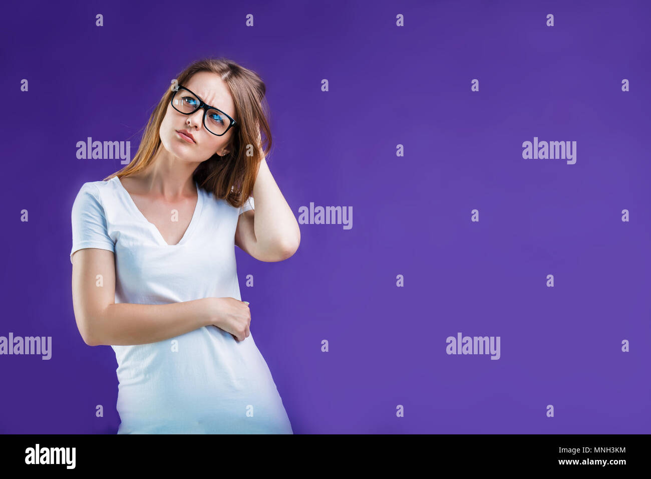 Portrait of pretty blonde woman dressed in white t-shirt and eyeglasses while thinking, isolated on blue studio background Stock Photo