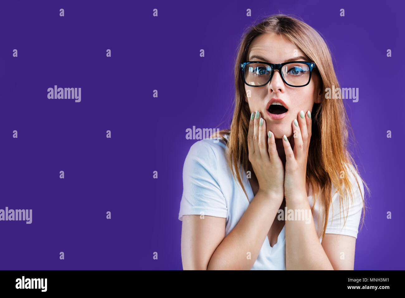 Portrait of pretty blonde woman dressed in white t-shirt and eyeglasses posing with excited face expression, isolated on blue studio background Stock Photo
