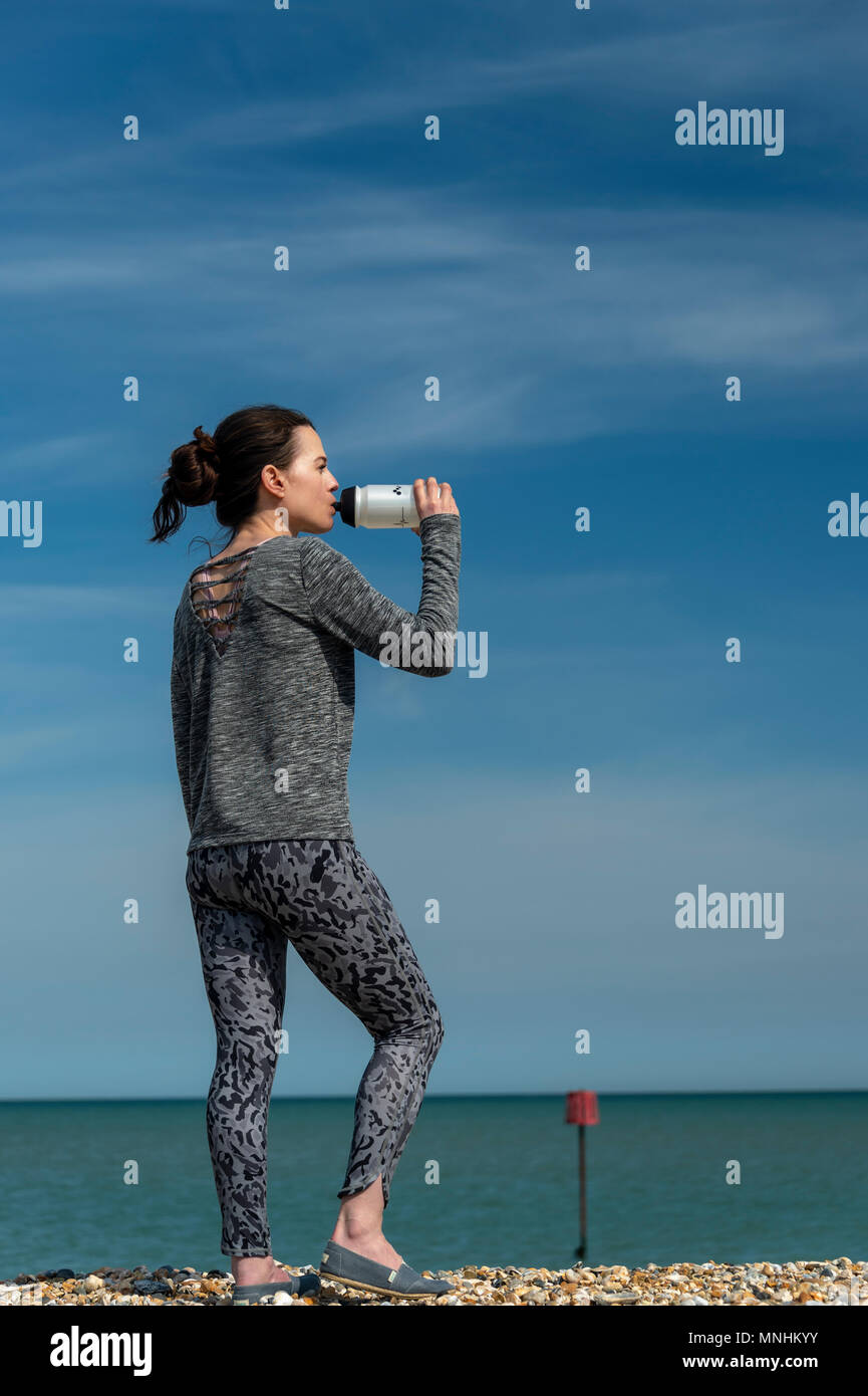 sporty woman standing by the sea resting and drinking from a sports water bottle Stock Photo