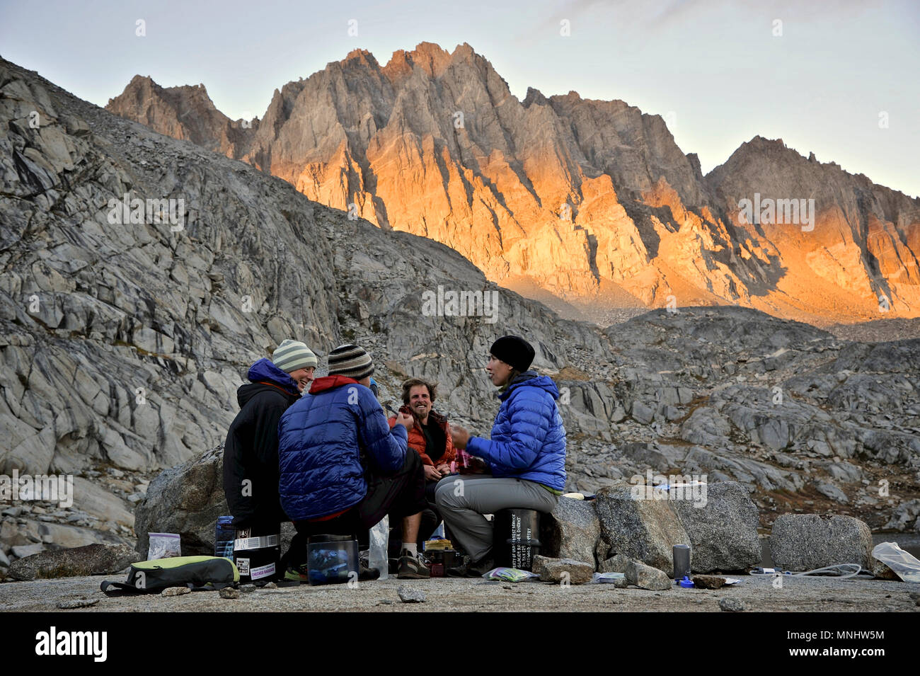 Backpackers have dinner with headlamps sitting at camp on the Barrett Lakes in Palisade Basin on a two-week trek of the Sierra High Route in Kings Canyon National Park in California. The 200-mile route roughly parallels the popular John Muir Trail through the Sierra Nevada Range of California from Kings Canyon National Park to Yosemite National Park. Sunset lights up the North Palisade in the background. Stock Photo