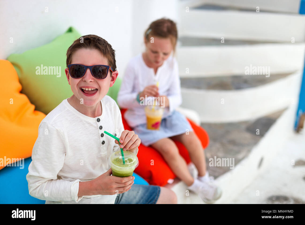 Two cute kids brother and sister drinking fresh smoothies on a colorful pillows at outdoor cafe on summer day Stock Photo