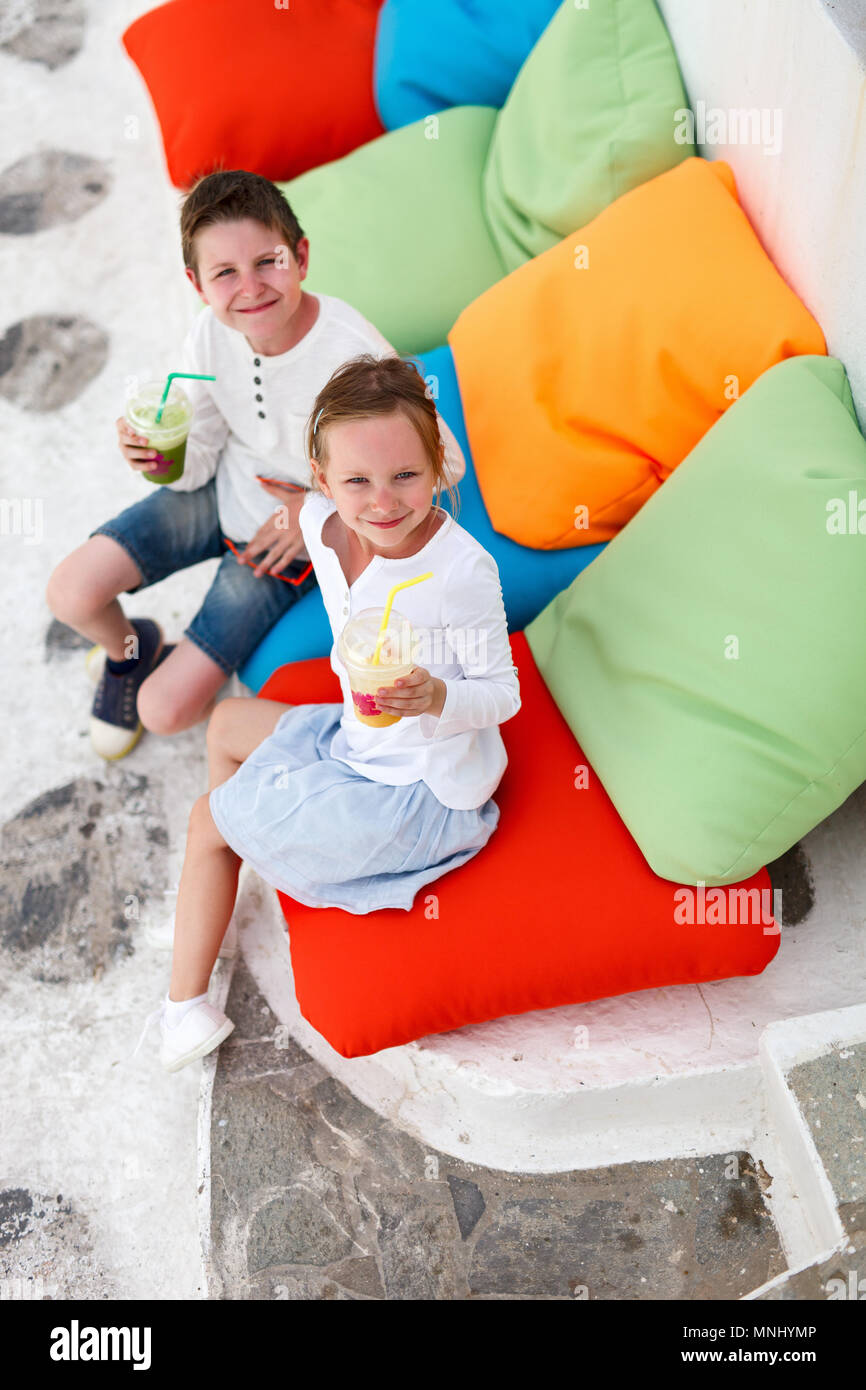 Two cute kids brother and sister drinking fresh smoothies on a colorful pillows at outdoor cafe on summer day Stock Photo