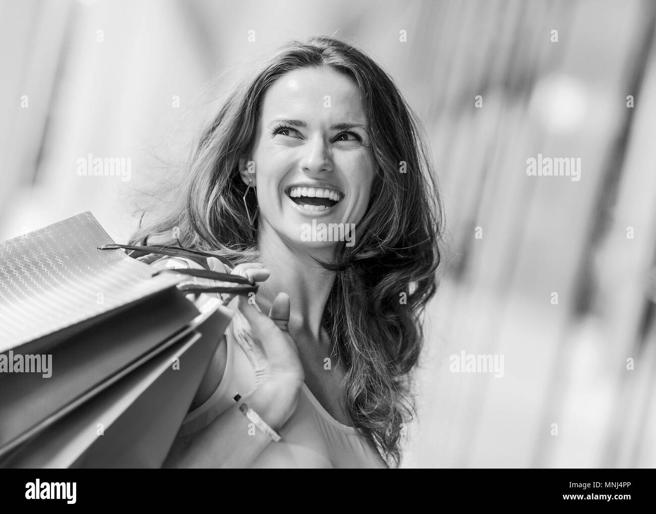 A brown-haired woman holding three shopping bags - brown, gold, and - over her right shoulder looks back at someone who is making her laugh. A good sh Stock Photo