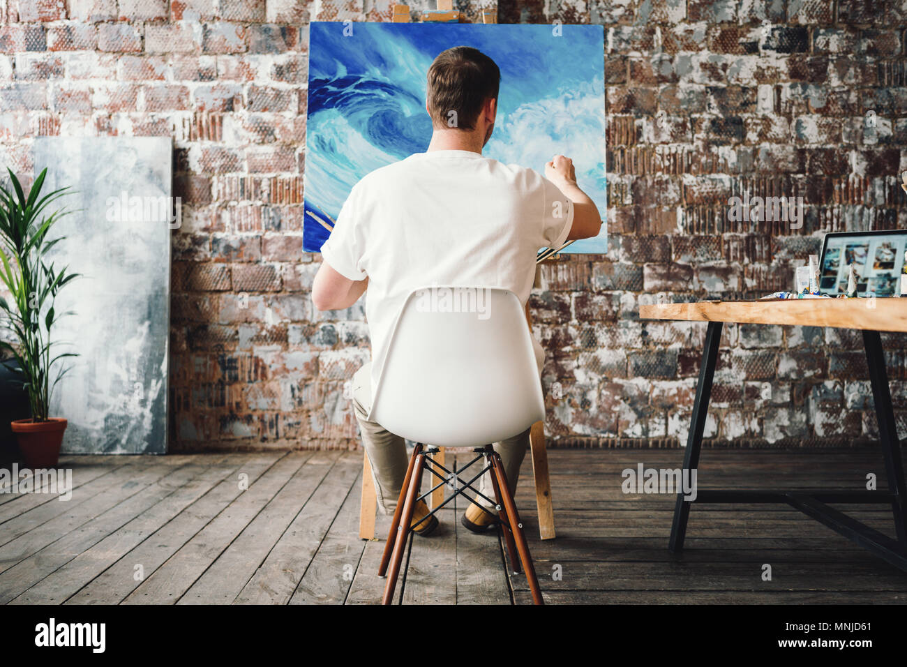 Professional painter sits on chair in front of easel with canvas, hold ...