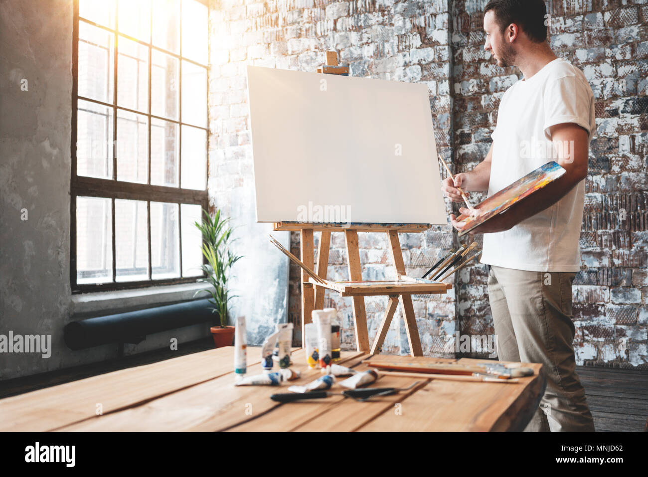 Man artist stands in front of a blank canvas on easel with palette in