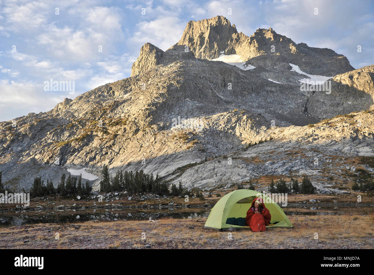 Backpacker in her sleeping bag on cold morning at camp near Thousand Island Lake with Banner Peak in background on trek of Sierra High Route in Minarets Wilderness, Inyo National Forest, California, USA Stock Photo