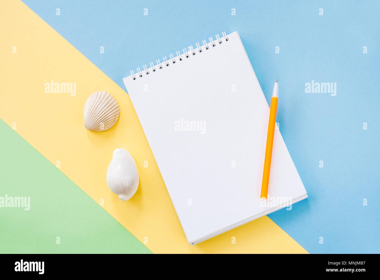 Summer vacations concept with yellow stripe, white notepad, orange pen and two seashells on blue-green background. Stock Photo