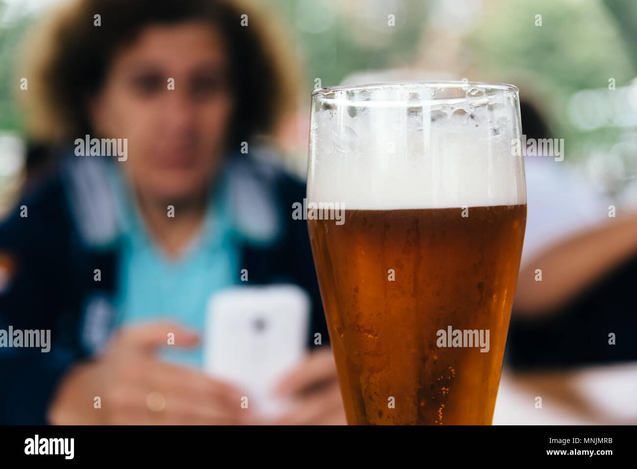 Close up of refreshing pint of lager beer against woman texting on smart phone. Selective focus Stock Photo