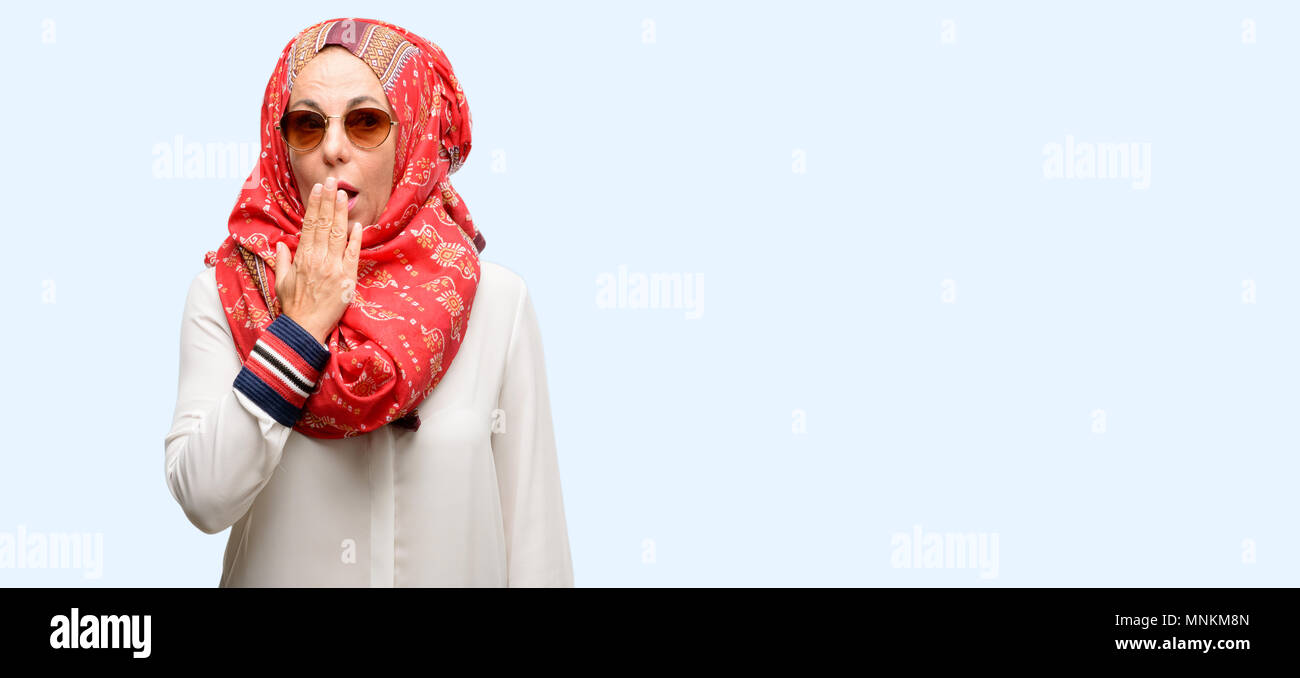 Middle age muslim arab woman wearing hijab covers mouth in shock, looks shy, expressing silence and mistake concepts, scared isolated blue background Stock Photo