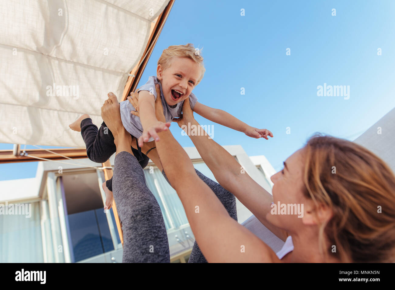 Happy mother playing with little boy at home. Woman holding her son on her legs while lying on sofa in doors. Stock Photo