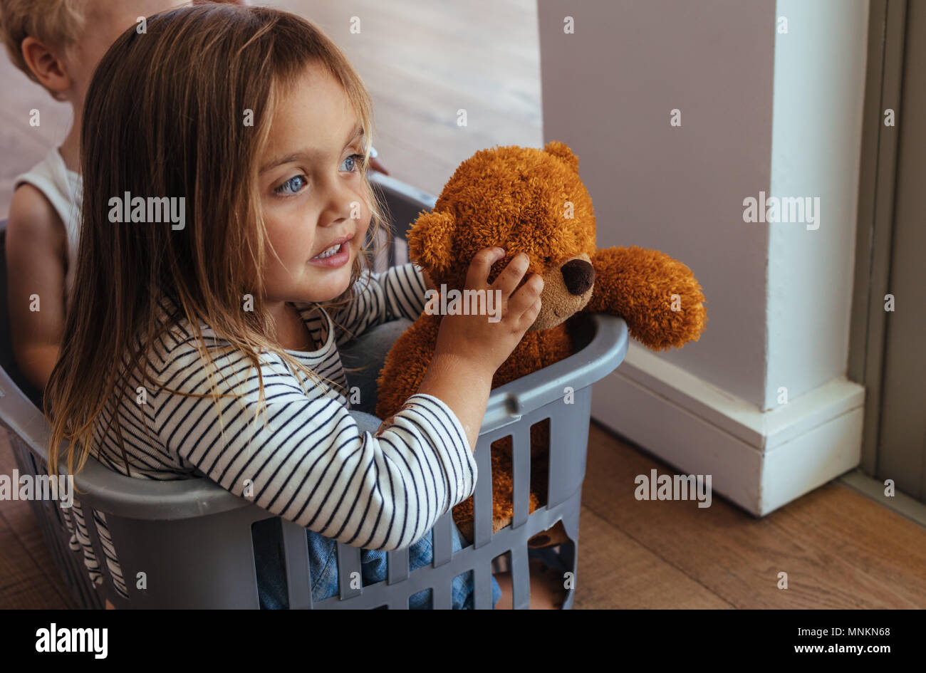 Beautiful little girl sitting in a washing basket with her teddy bear and brother at the back. Siblings playing in a washing basket at home. Stock Photo