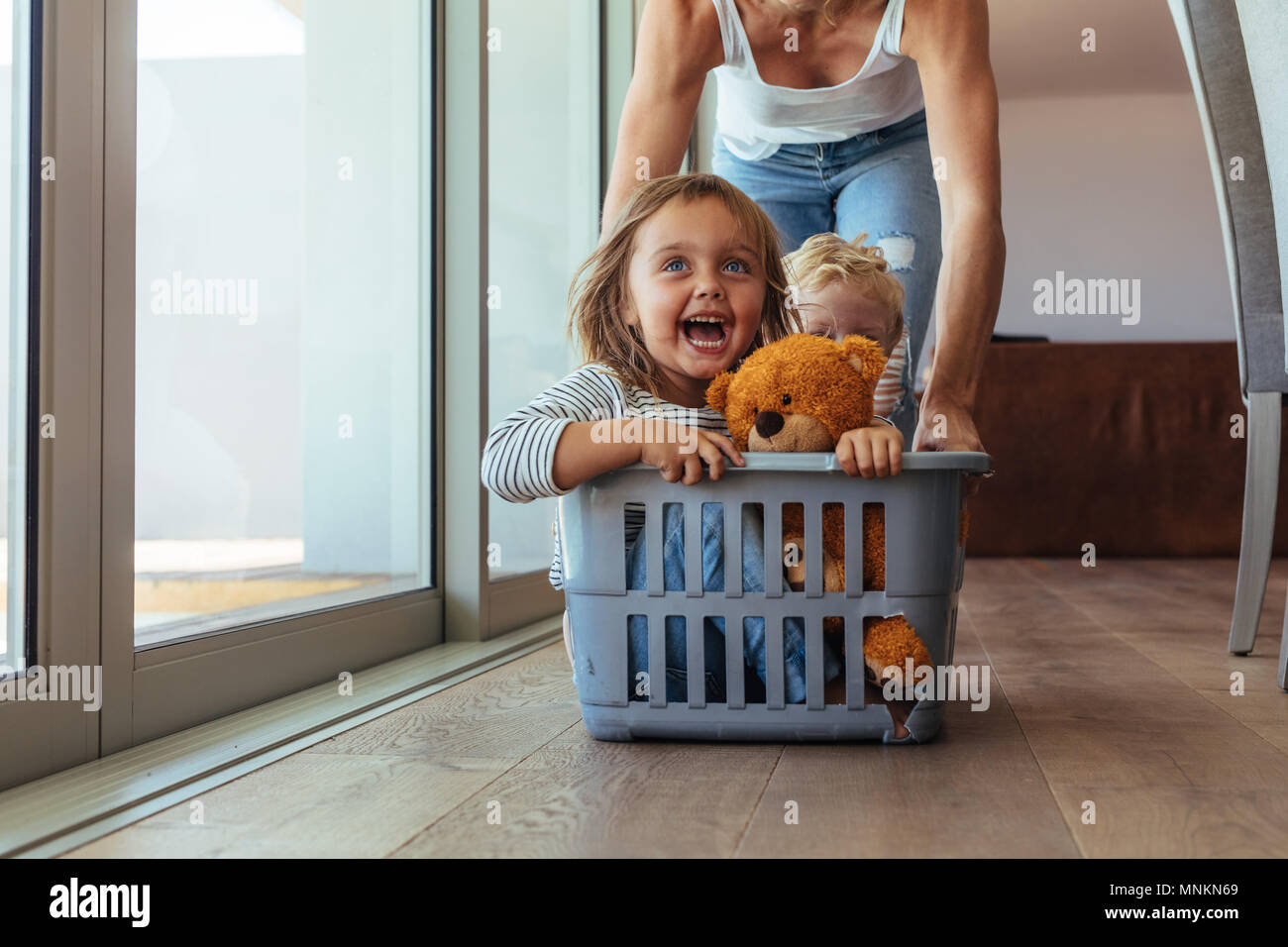 Excited little girl with teddy bear and her brother sitting in a laundry basket being pushed by her mother. Cute family playing together at home. Stock Photo