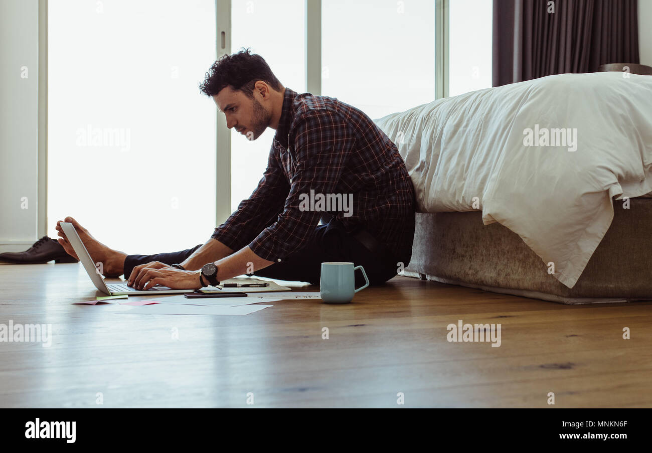 Man working on laptop computer sitting on the floor. Freelancer sitting on floor beside a bed and working on laptop at home with a coffee cup by his s Stock Photo
