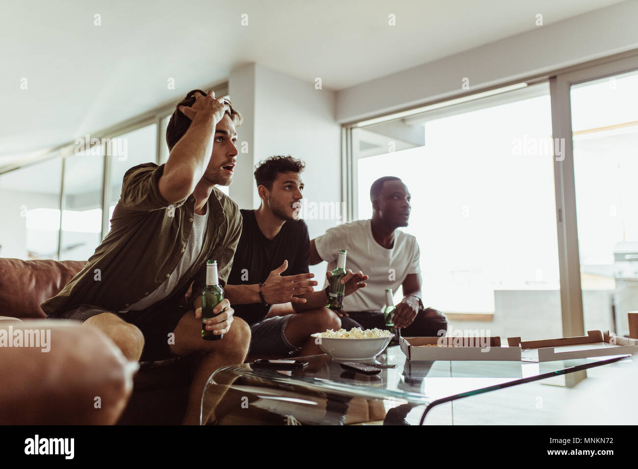 Three friends watching a game on television holding beer bottles. Men sitting on sofa with drinks and snacks watching television at home. Stock Photo