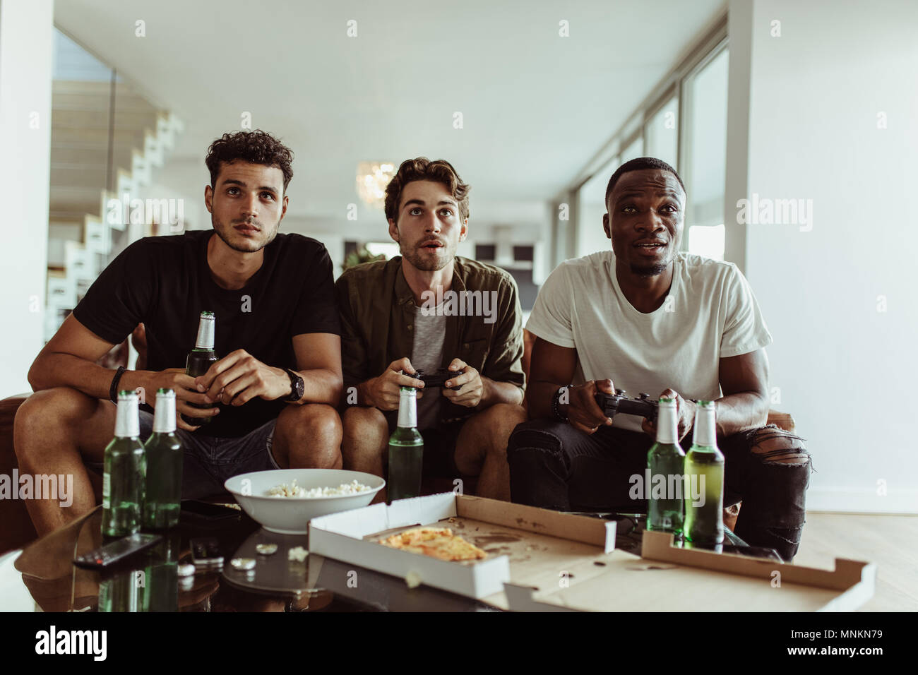 Friends having fun with drinks and snacks while playing video game at home. Two men playing video game holding joysticks while another man watches wit Stock Photo