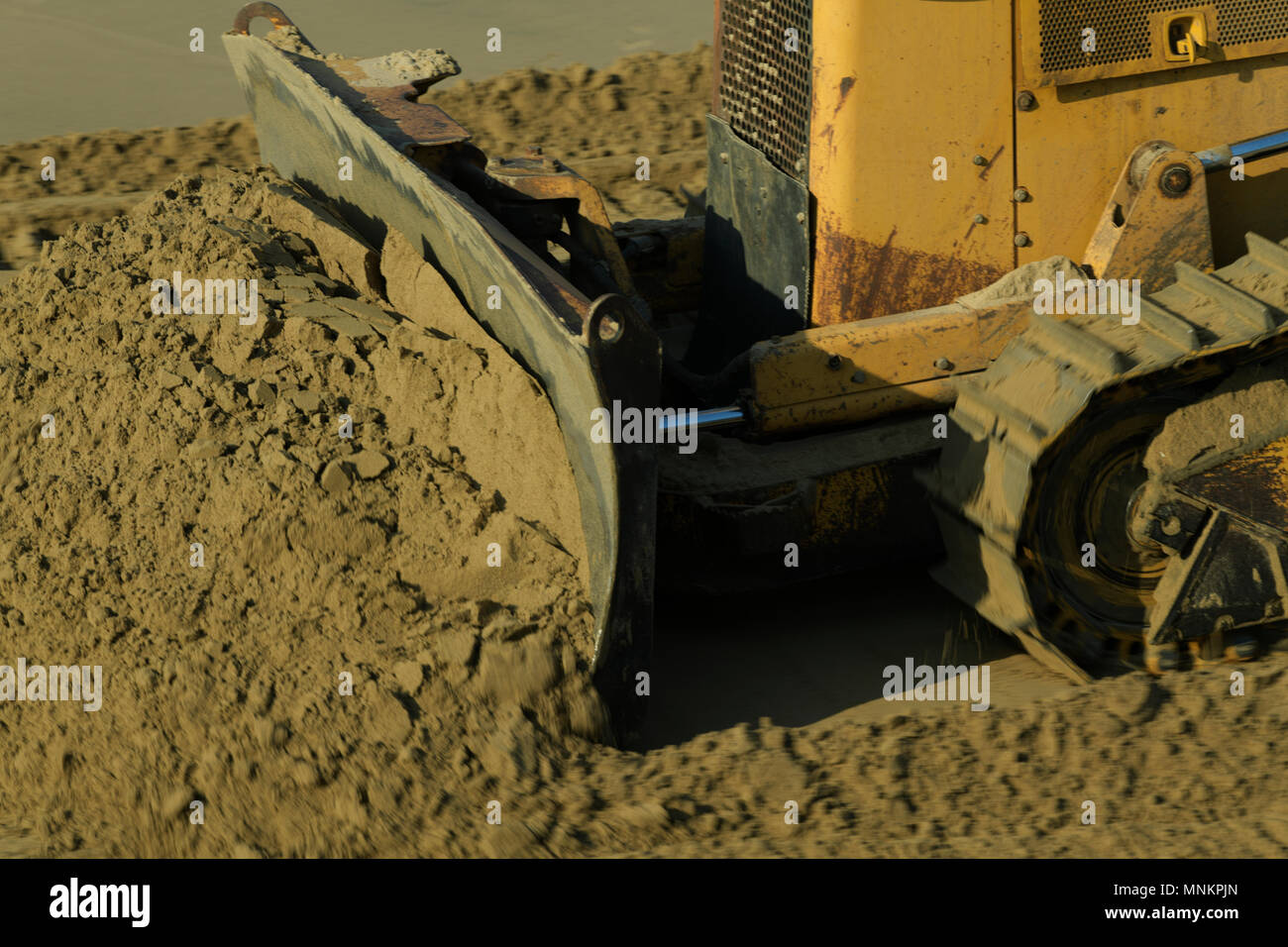 Close-up, detail, blur, scraper blade of old yellow construction bulldozer pushing crumbling wet sand, motion Stock Photo