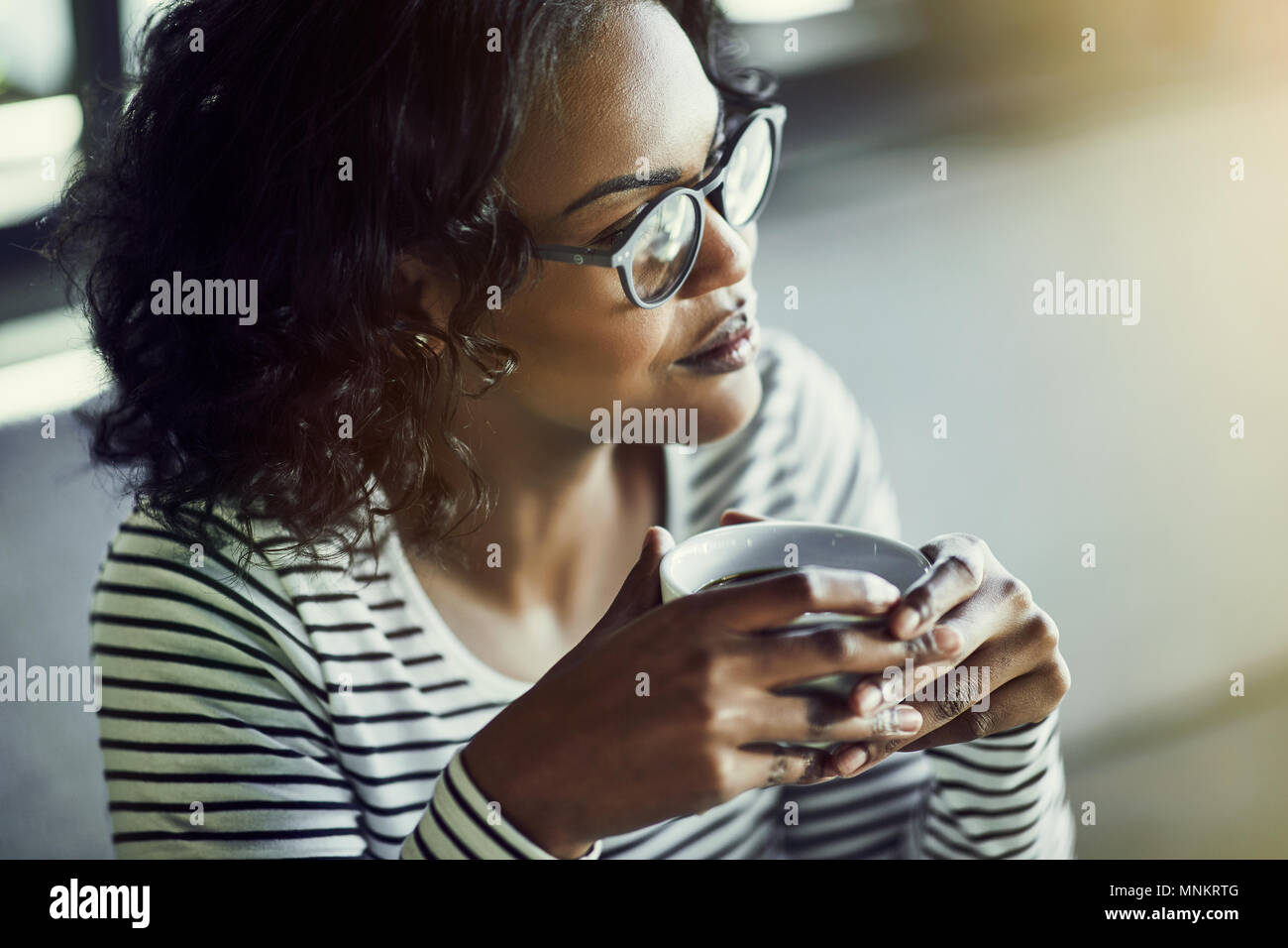 Smiling young African woman wearing glasses sitting alone at a cafe table deep in thought while drinking a fresh cup of coffee Stock Photo