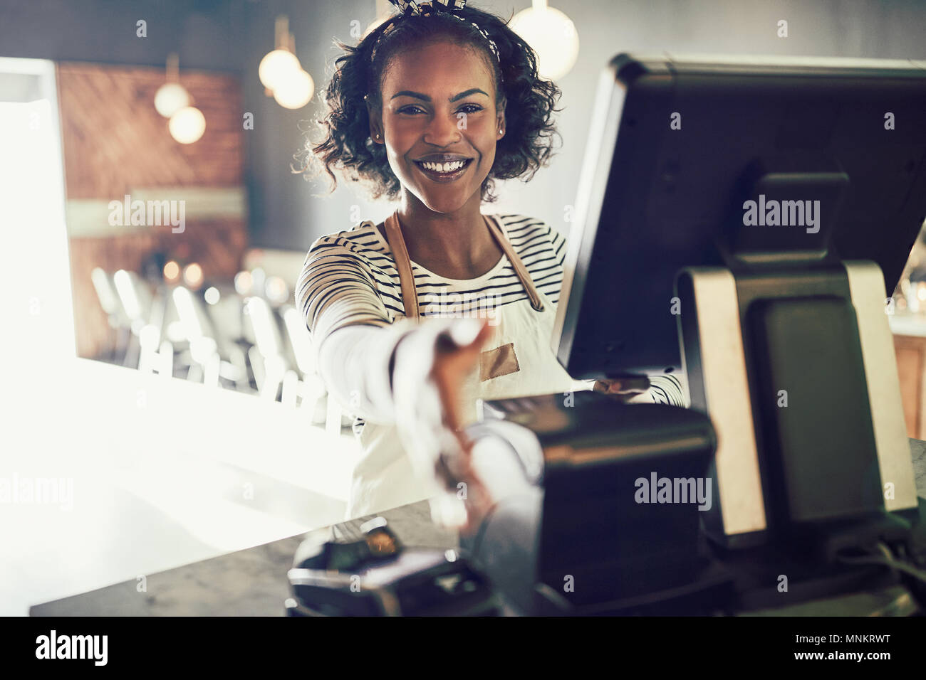 Smiling young African cafe owner standing at a point of sale terminal in her restaurant extending a handshake Stock Photo