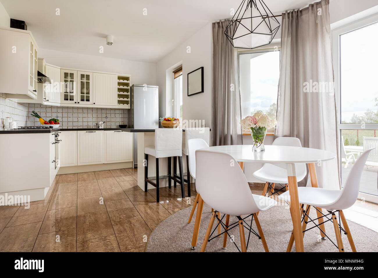 Light stylish interior with round table, white chairs and functional open kitchen Stock Photo