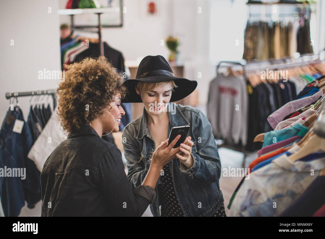 Millennial female friends looking at a smartphone in a vintage clothing store Stock Photo