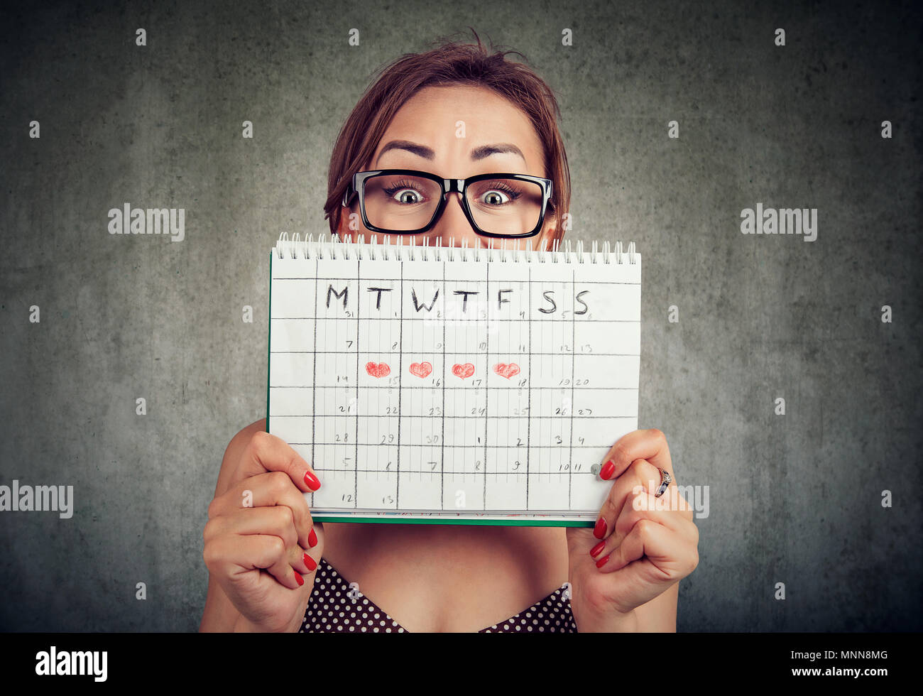 Portrait of a funny young woman in glasses hiding behind a periods calendar and looking at camera isolated gray wall background Stock Photo